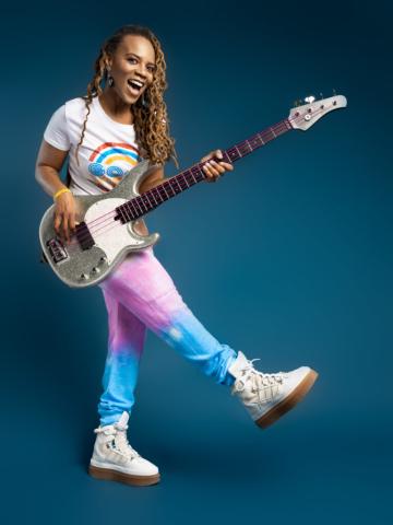 Divinity Roxx holding guitar, wearing a rainbow shirt with one leg raised.